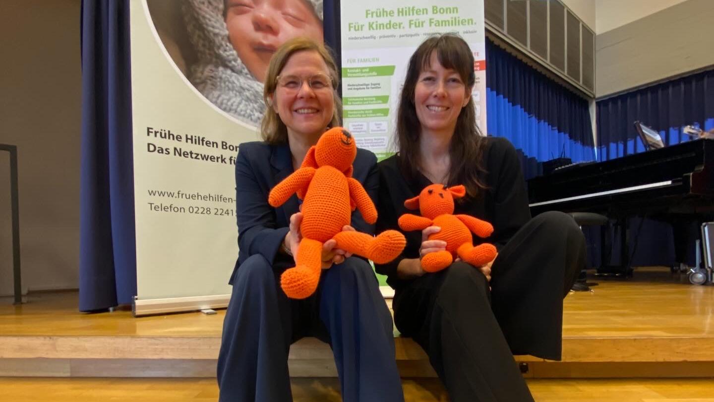 Die Leiterinnen der Koordinierungsgruppe Frühe Hilfen Anja Henkel und Susanne Absalon sitzen auf der Treppe im Veranstaltungsraum gemeinsam mit den beiden orangenen Maskottchen der Frühen Hilfen