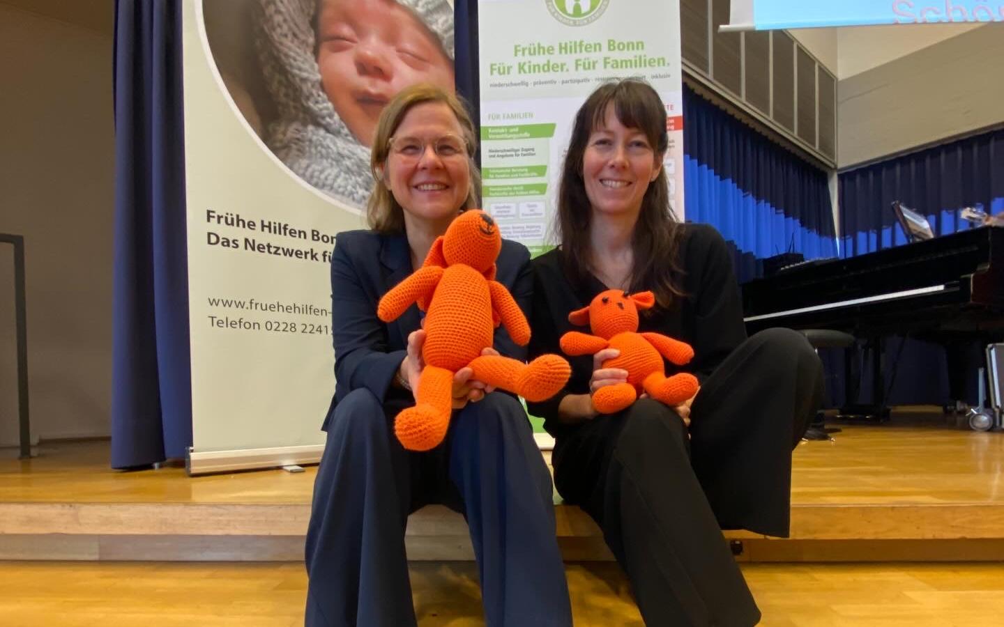 Die Leiterinnen der Koordinierungsgruppe Frühe Hilfen Anja Henkel und Susanne Absalon sitzen auf der Treppe im Veranstaltungsraum gemeinsam mit den beiden orangenen Maskottchen der Frühen Hilfen