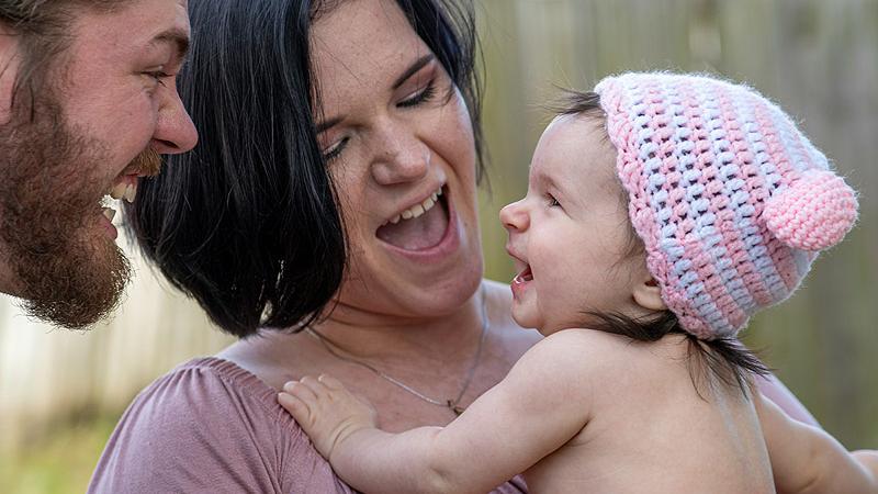 Herzhaft lachende Mutter hält lachendes Baby auf dem Arm. Das Baby lacht den Vater an, der ebenfalls lacht. und