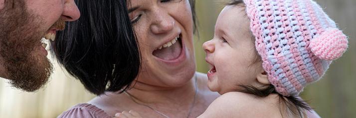 Herzhaft lachende Mutter hält lachendes Baby auf dem Arm. Das Baby lacht den Vater an, der ebenfalls lacht. und
