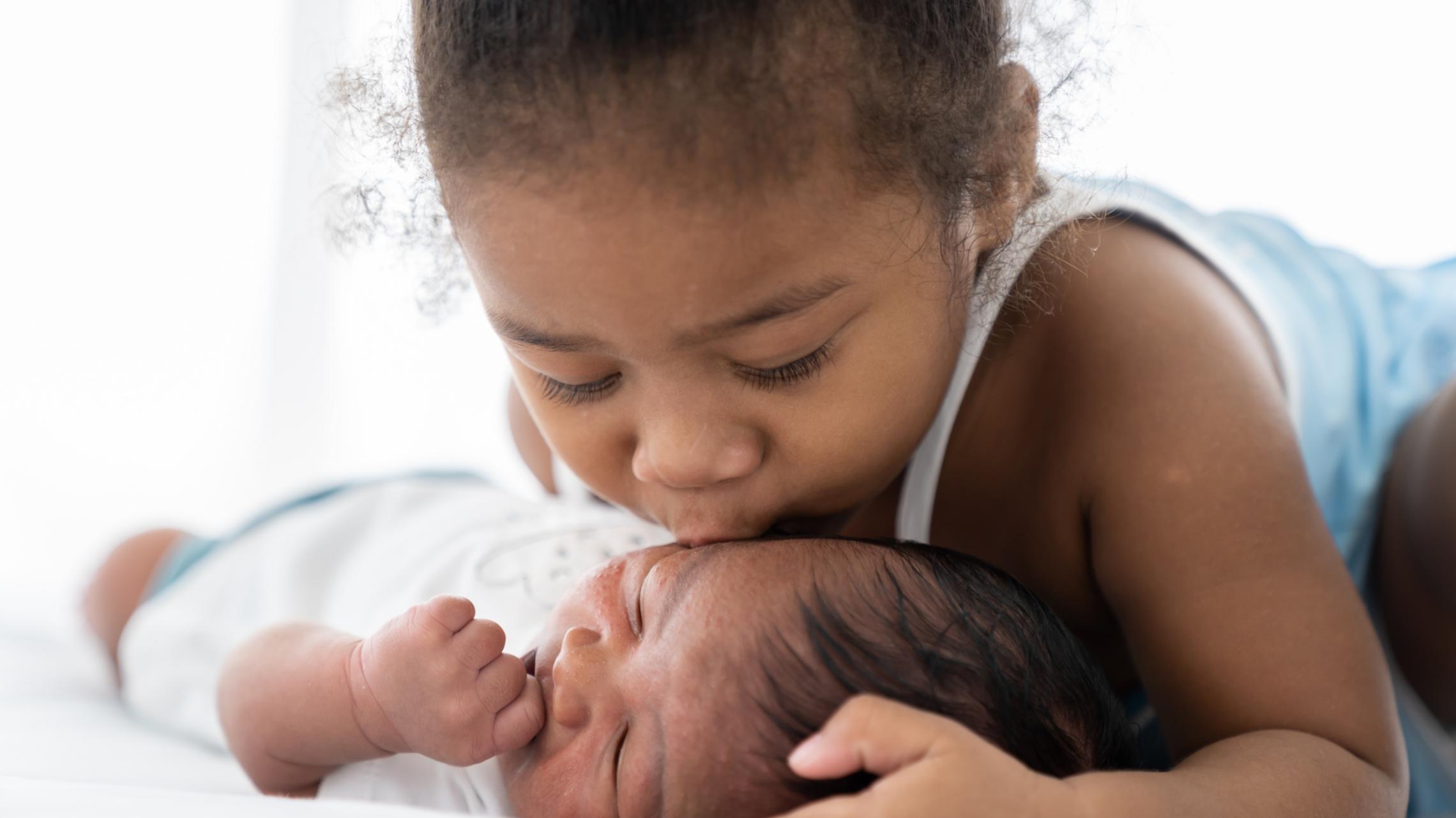 Kleinkind küsst zärtlich ein auf dem Boden liegendem Baby auf die Backe