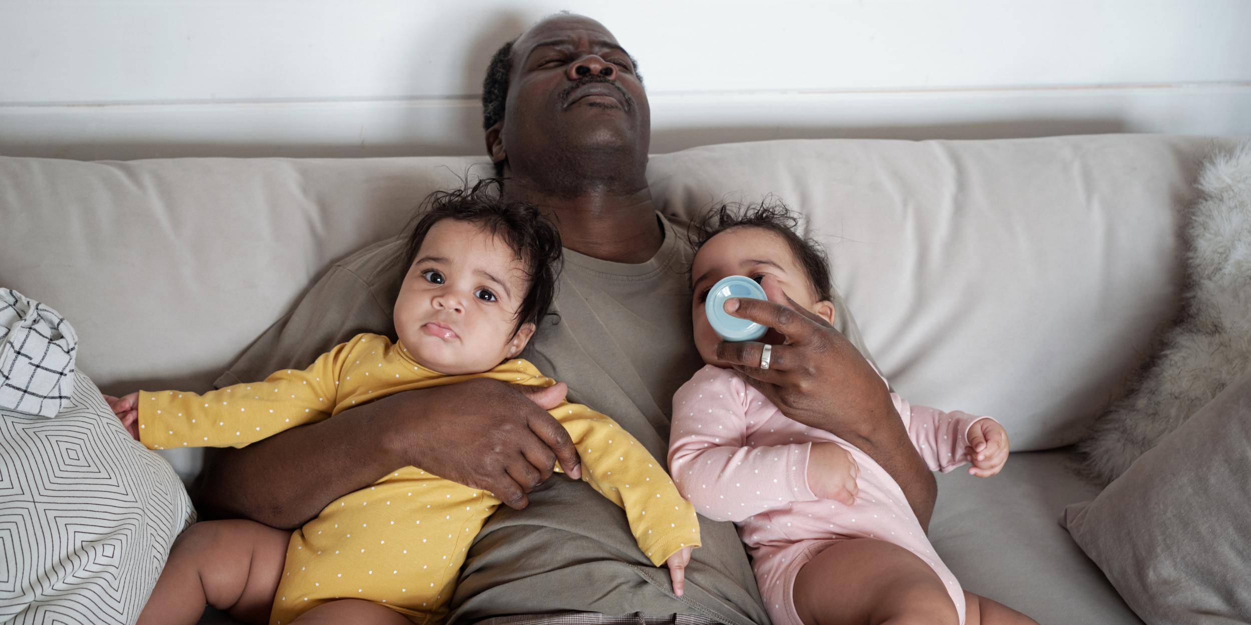 erschöpfter Vater auf der Couch mit 2 Babys im Arm und ein Baby trinkt aus der Flasche