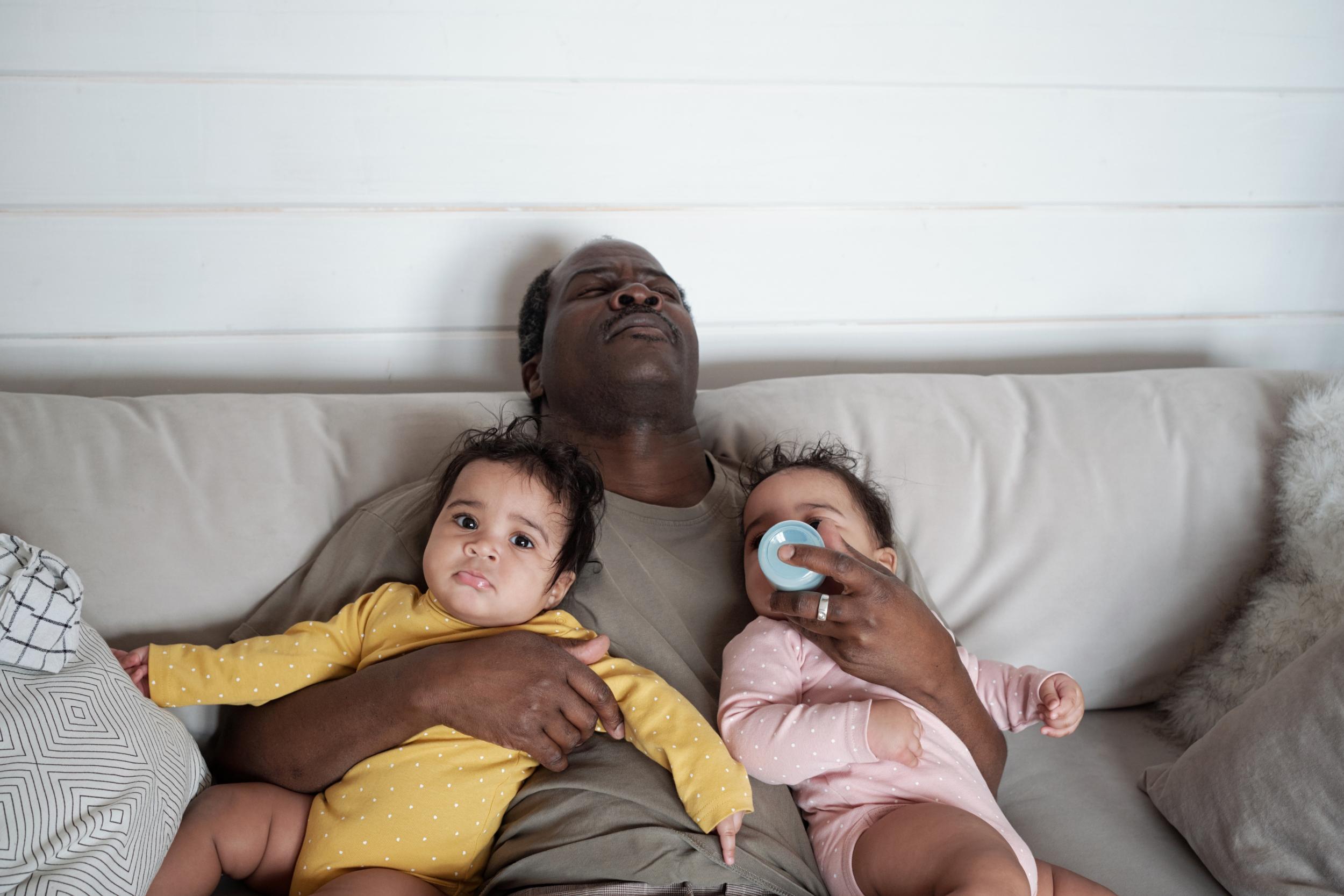 erschöpfter Vater auf der Couch mit 2 Babys im Arm und ein Baby trinkt aus der Flasche