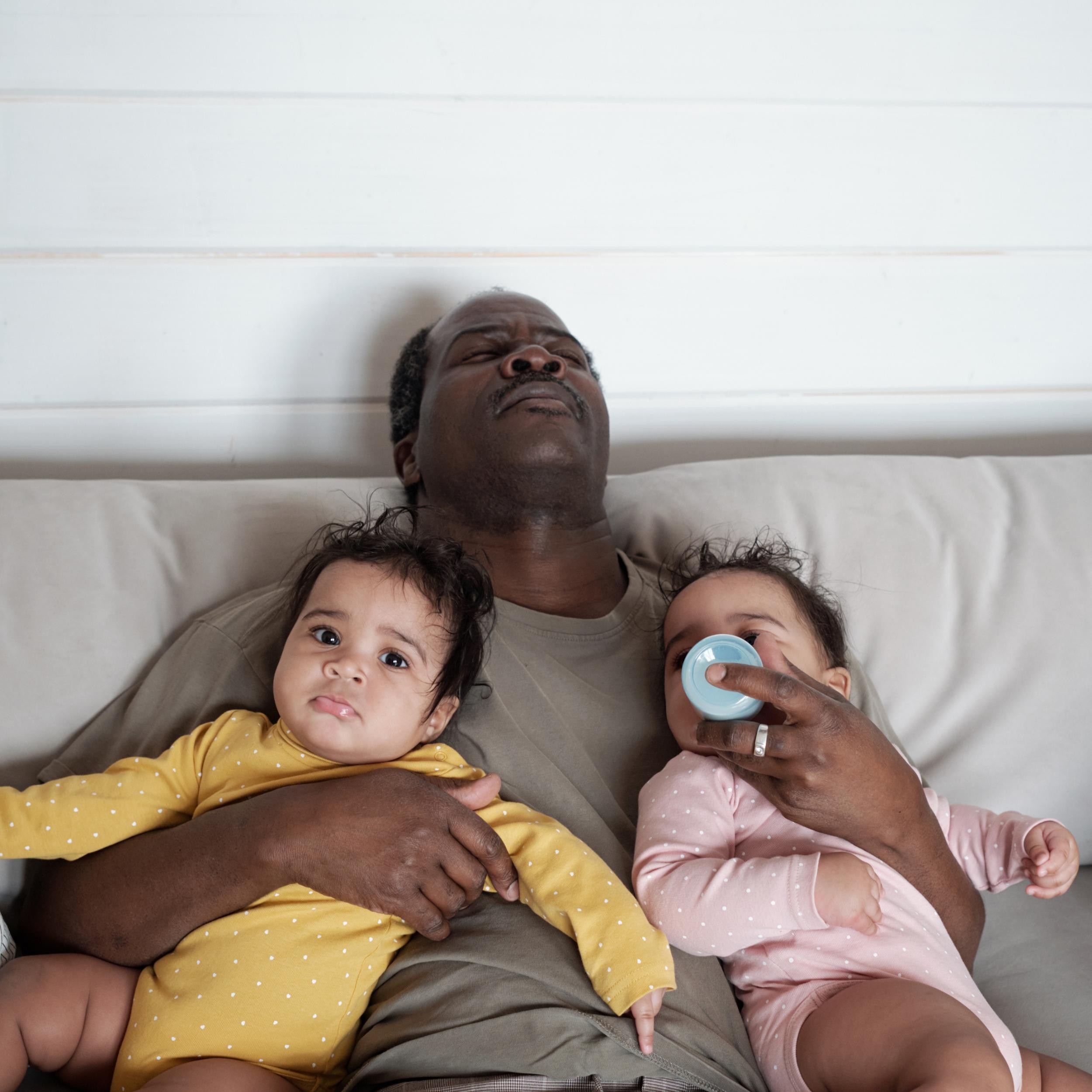 erschöpfter Vater auf der Couch mit 2 Babys im Arm und ein Baby trinkt aus der Flasche