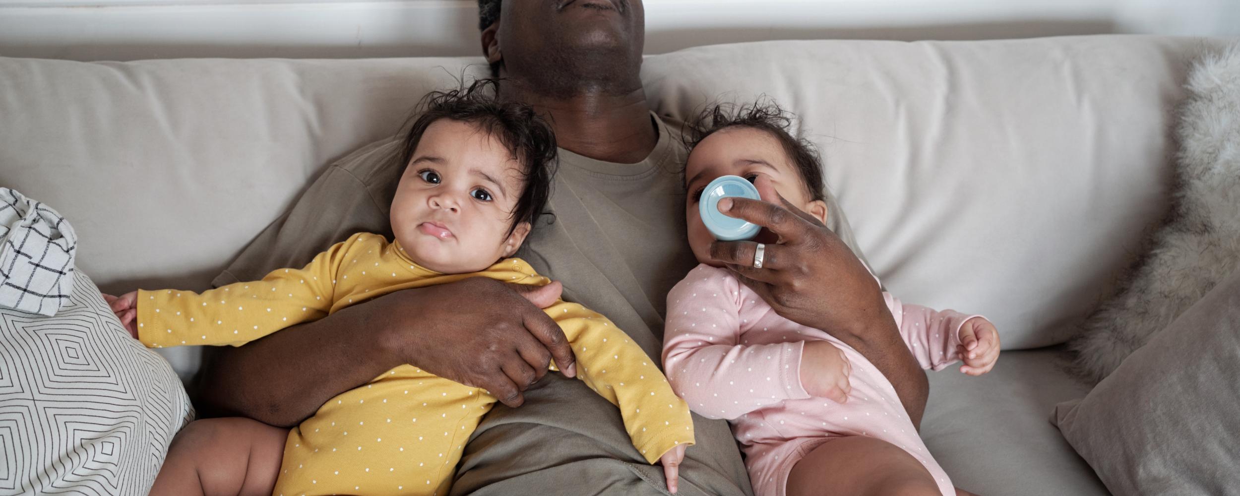 erschöpfter Vater auf der Couch mit 2 Babys im Arm und ein Baby trinkt aus der Flasche