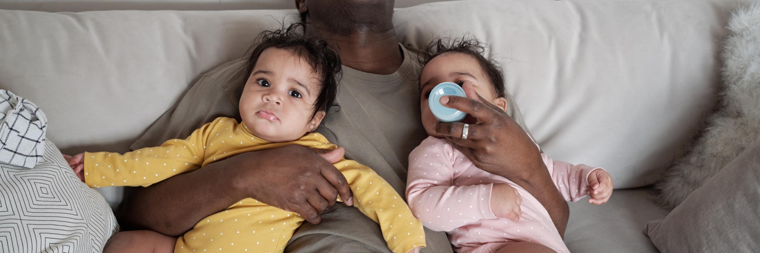 erschöpfter Vater auf der Couch mit 2 Babys im Arm und ein Baby trinkt aus der Flasche