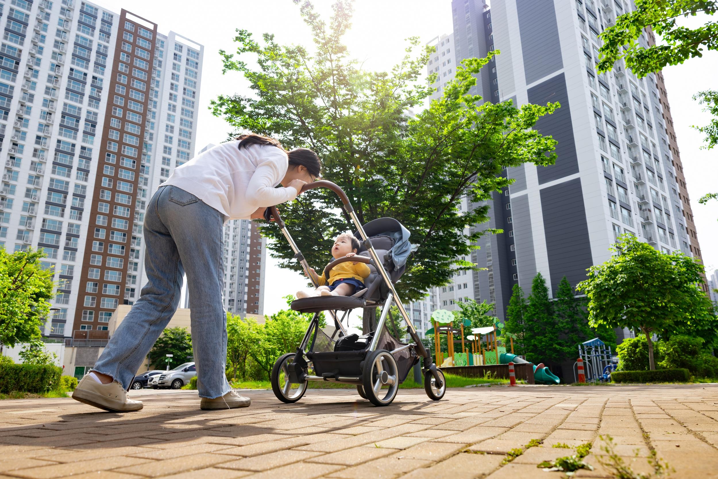 Mutter schiebt ein Kleinkind im Kinderwagen durch Straße in einer Großstadt. Im Hintergrund sind zwei Hochhäuser zu sehen.