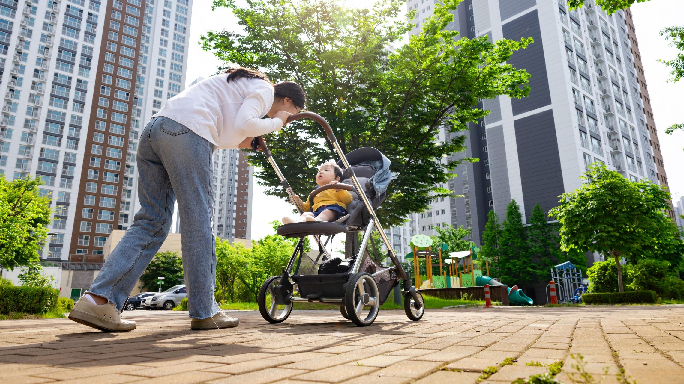 Mutter schiebt ein Kleinkind im Kinderwagen durch Straße in einer Großstadt. Im Hintergrund sind zwei Hochhäuser zu sehen.