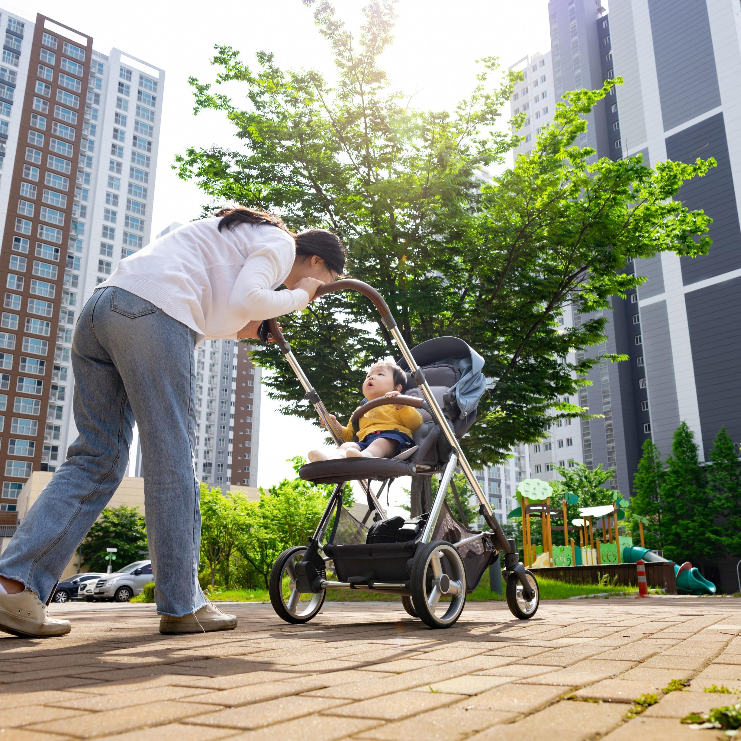Mutter schiebt ein Kleinkind im Kinderwagen durch Straße in einer Großstadt. Im Hintergrund sind zwei Hochhäuser zu sehen.