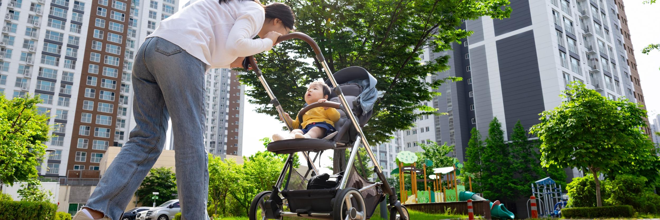 Mutter schiebt ein Kleinkind im Kinderwagen durch Straße in einer Großstadt. Im Hintergrund sind zwei Hochhäuser zu sehen.