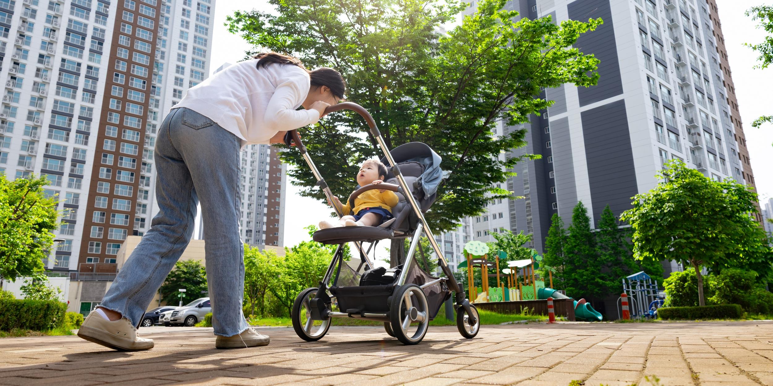 Mutter schiebt ein Kleinkind im Kinderwagen durch Straße in einer Großstadt. Im Hintergrund sind zwei Hochhäuser zu sehen.