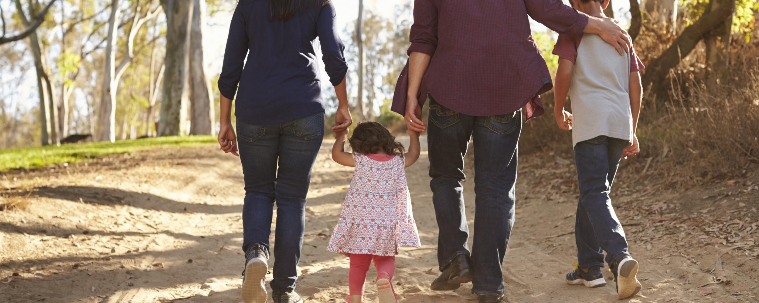 Vater, Mutter und 2 Kinder bei einem Spaziergang auf sandigem Boden und Wald. Alle 4 halten sich an der Hand und sind nur mit dem Rücken zu sehen