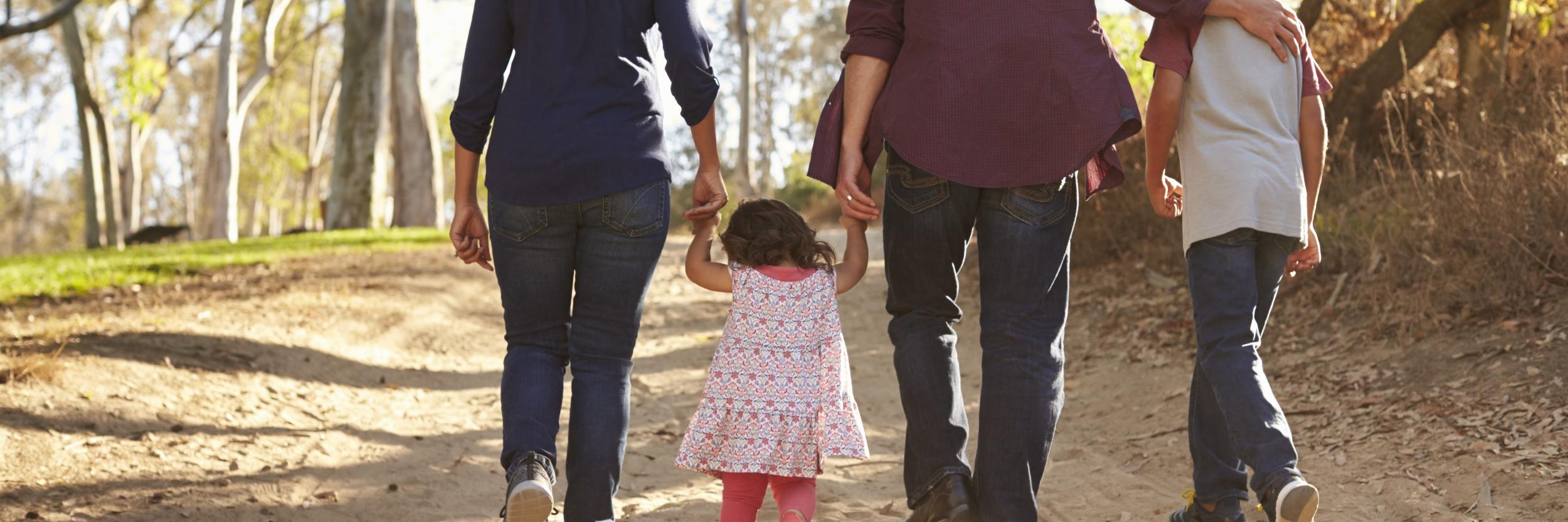Vater, Mutter und 2 Kinder bei einem Spaziergang auf sandigem Boden und Wald. Alle 4 halten sich an der Hand und sind nur mit dem Rücken zu sehen
