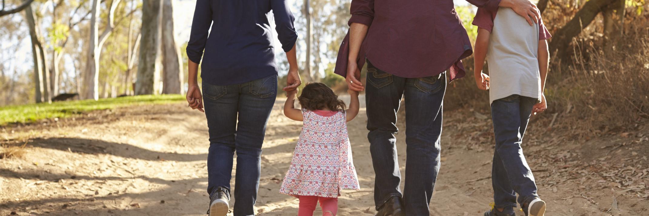 Vater, Mutter und 2 Kinder bei einem Spaziergang auf sandigem Boden und Wald. Alle 4 halten sich an der Hand und sind nur mit dem Rücken zu sehen