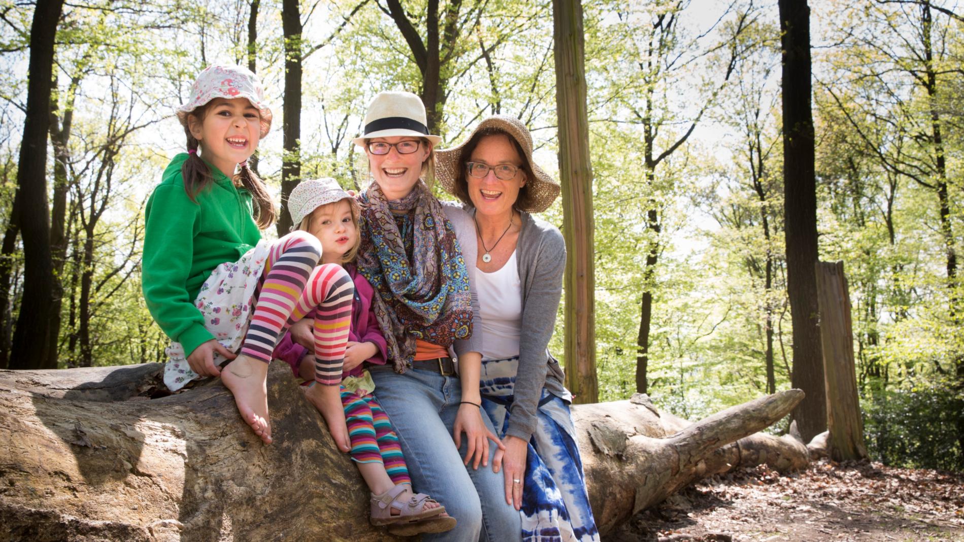 Mütter mit Kindern im Wald. Alle tragen einen Hut und sitzen auf einem Baumstamm.