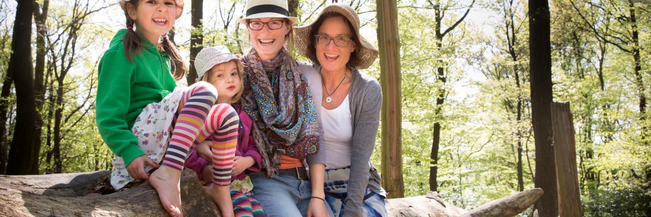 Mütter mit Kindern im Wald. Alle tragen einen Hut und sitzen auf einem Baumstamm.