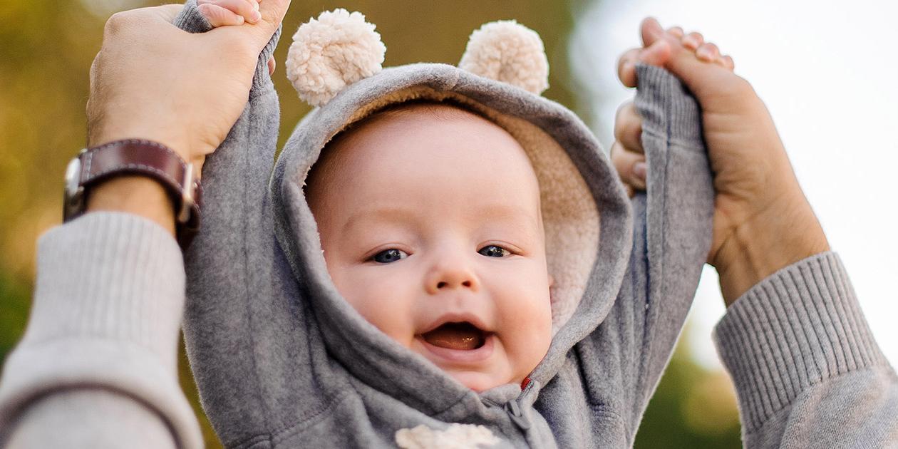 Bild mit lächelndem Baby, das die Arme mit Hilfe eines Elternteils nach oben streckt.