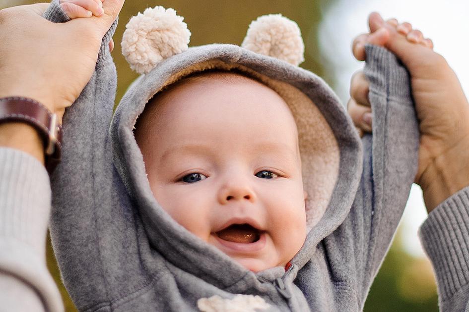 Bild mit lächelndem Baby, das die Arme mit Hilfe eines Elternteils nach oben streckt.