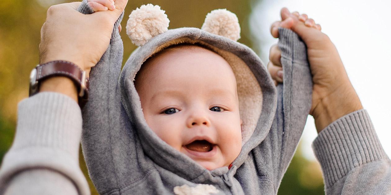 Bild mit lächelndem Baby, das die Arme mit Hilfe eines Elternteils nach oben streckt.