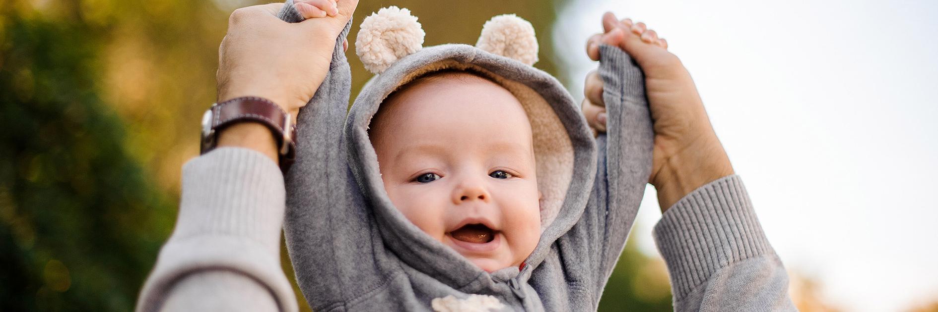 Bild mit lächelndem Baby, das die Arme mit Hilfe eines Elternteils nach oben streckt.